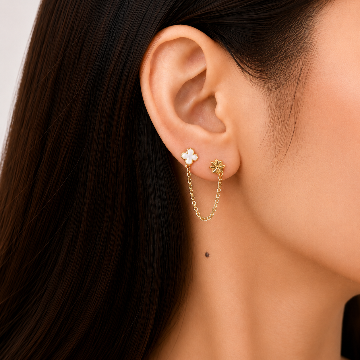 Close-up of a woman wearing gold-tone double piercing earrings featuring a white clover stud and star stud connected by a fine chain.